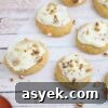25+ Dairy-Free, Gluten-Free Protein Desserts 17 Dairy-free collagen carrot cookies topped with cream cheese frosting and pecans on a piece of parchment paper with shredded carrots beside them.