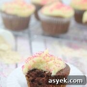 25+ Dairy-Free, Gluten-Free Protein Desserts 20 Chocolate protein cupcake with buttercream frosting with a bite taken out of it on a white chupcake liner with more gluten-free cupcakes on a cooling rack behind it.