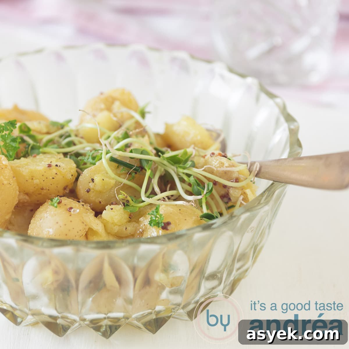a square photo of a glass bowl filled with potato salad and some greens.